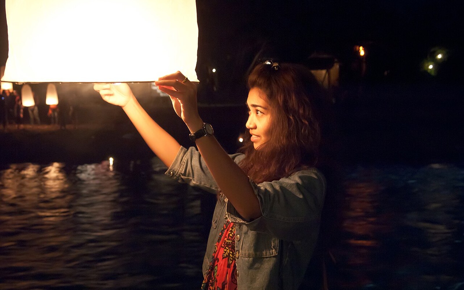Woman lifting a glowing sky lantern beside water at night