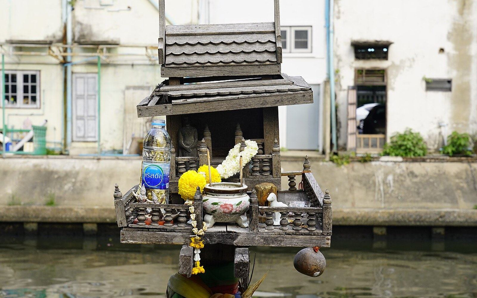 Wooden spirit house with offerings and incense pot beside a canal